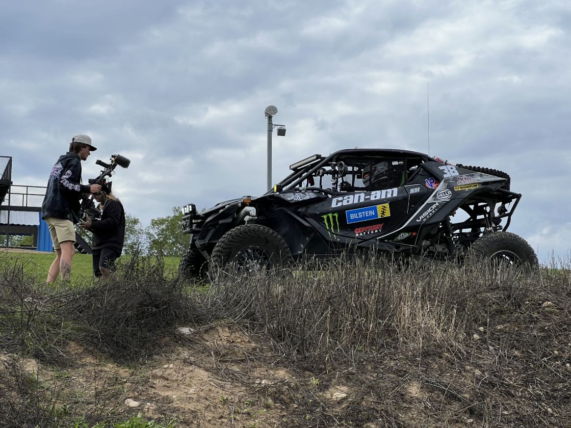 Camera operator with a gimbal rig filming a side-by-side off-road race vehicle on a grassy hilltop