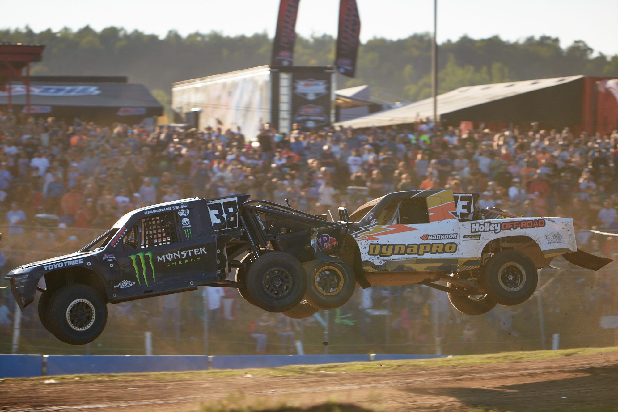 Two pro short-course trucks side by side in the air over a jump, packed grandstand behind