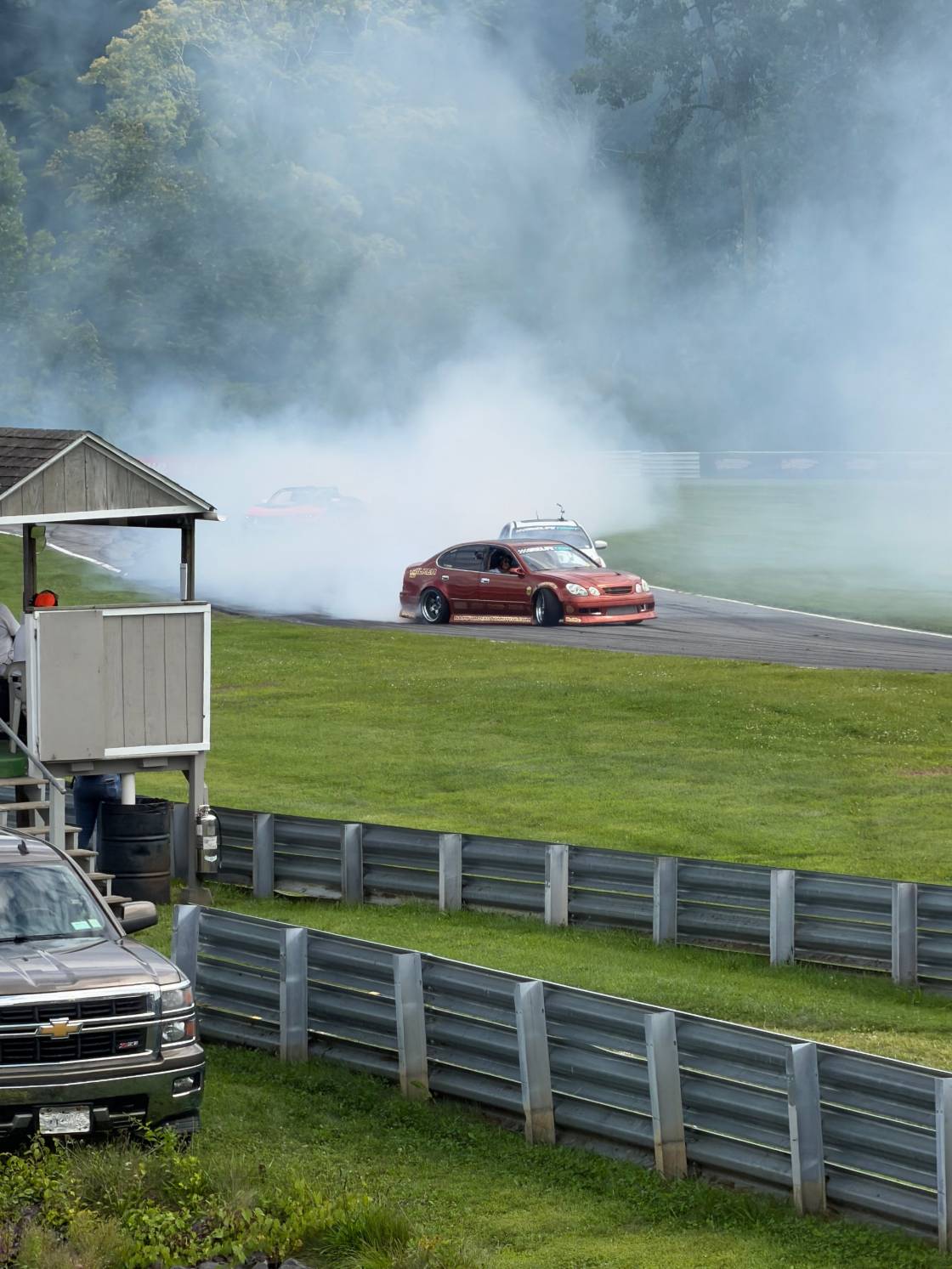 Red sedan drifting through a cloud of tire smoke on a tree-lined track at GRIDLIFE