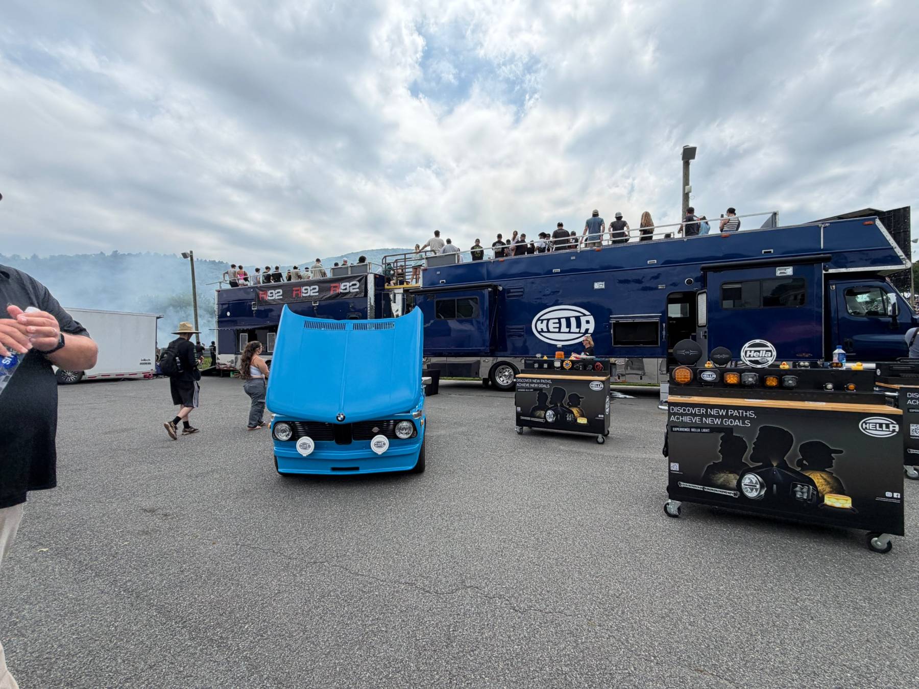 Light blue vintage sedan with its hood up and auxiliary driving lights, in front of a double-deck hospitality rig with fans on the roof deck at GRIDLIFE