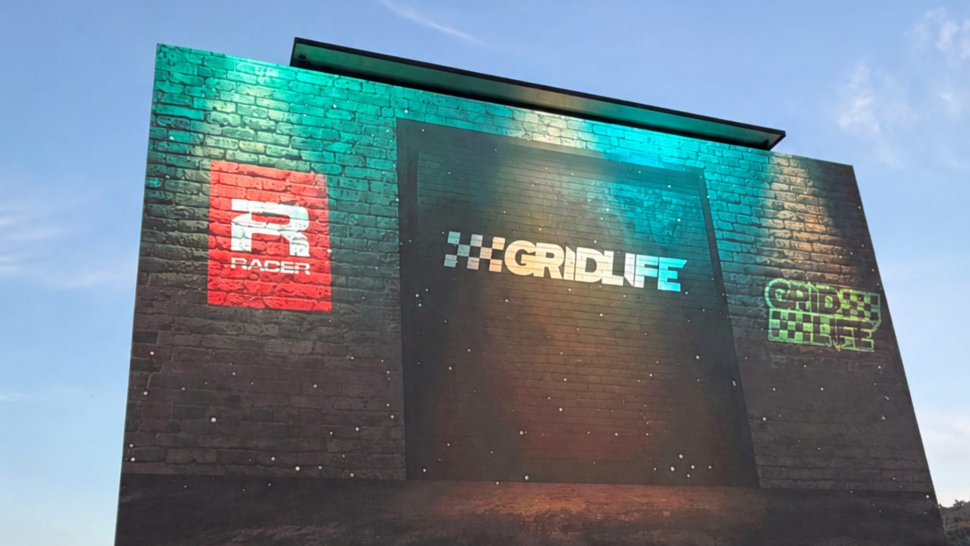 GRIDLIFE logo lit in teal and gold on a brick-textured screen wall against a blue evening sky
